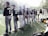 Players in baseball uniforms stand in a line on a field, facing away. Most are wearing dark blue jerseys with numbers and names on the back. Hands holding gloves are visible, and various equipment is scattered around them. The field is surrounded by a fence, and trees can be seen in the background.