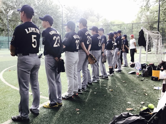 Players in baseball uniforms stand in a line on a field, facing away. Most are wearing dark blue jerseys with numbers and names on the back. Hands holding gloves are visible, and various equipment is scattered around them. The field is surrounded by a fence, and trees can be seen in the background.