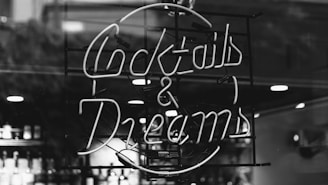 A black and white image featuring a neon sign with the words 'Cocktails & Dreams.' The sign is illuminated and likely situated in a bar or lounge setting, with faint outlines of bottles and lights visible in the background.