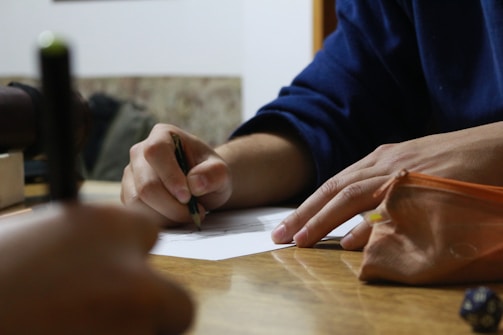 Two hands are engaged in a writing activity, with one holding a pencil over a piece of paper. A pencil case rests on the table nearby, and a blurred object is visible in the foreground. The setting appears to be a casual workspace.