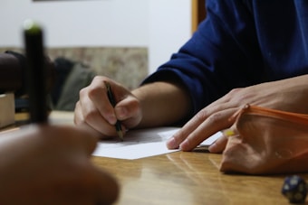 Two hands are engaged in a writing activity, with one holding a pencil over a piece of paper. A pencil case rests on the table nearby, and a blurred object is visible in the foreground. The setting appears to be a casual workspace.