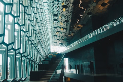 A modern architectural interior with a striking geometric design. Large windows with a hexagonal pattern allow natural light to flood the space, while a staircase ascends alongside the wall. The ceiling features reflective, metallic tiles that enhance the contemporary aesthetic.