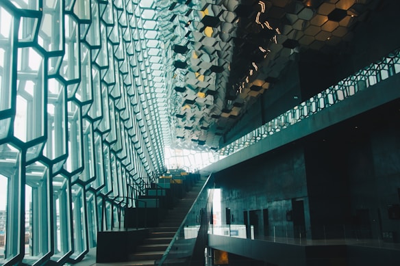 A modern architectural interior with a striking geometric design. Large windows with a hexagonal pattern allow natural light to flood the space, while a staircase ascends alongside the wall. The ceiling features reflective, metallic tiles that enhance the contemporary aesthetic.