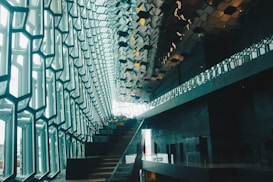 A modern architectural interior with a striking geometric design. Large windows with a hexagonal pattern allow natural light to flood the space, while a staircase ascends alongside the wall. The ceiling features reflective, metallic tiles that enhance the contemporary aesthetic.