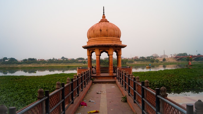 A traditional Indian pavilion with a domed roof made of reddish-brown stone stands on a narrow path leading into a calm river. The pavilion is surrounded by lush greenery and water plants. In the distance, a bridge stretches across the river, with buildings visible beyond, suggesting an urban setting. The scene appears peaceful and serene.