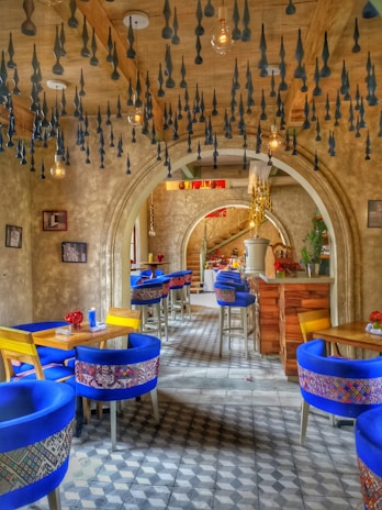 An artistic and vibrant café interior with blue chairs featuring patterned fabric, wooden tables, and decorative ceiling fixtures. The room features a series of arches and a checkered tiled floor, creating a cozy but stylish atmosphere.