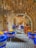 An artistic and vibrant café interior with blue chairs featuring patterned fabric, wooden tables, and decorative ceiling fixtures. The room features a series of arches and a checkered tiled floor, creating a cozy but stylish atmosphere.
