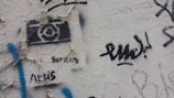 A textured wall features various pieces of street art and graffiti. There is a black stencil of a camera with text beneath it, which is partially obscured by peeling wall paint. Blue, black, and other colored graffiti marks are scattered around, adding a sense of urban artistic expression.