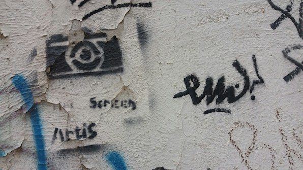 A textured wall features various pieces of street art and graffiti. There is a black stencil of a camera with text beneath it, which is partially obscured by peeling wall paint. Blue, black, and other colored graffiti marks are scattered around, adding a sense of urban artistic expression.