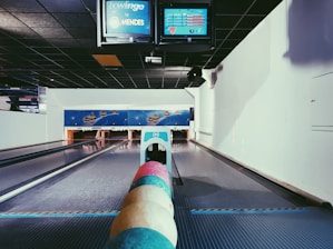 A bowling alley features multiple pastel-colored bowling balls aligned in the foreground. The alley extends towards the pins, with digital screens overhead displaying scores and information. The surrounding walls and ceiling are primarily dark and white, with a space-themed mural on the back wall.