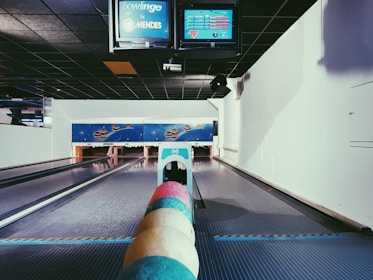 A bowling alley features multiple pastel-colored bowling balls aligned in the foreground. The alley extends towards the pins, with digital screens overhead displaying scores and information. The surrounding walls and ceiling are primarily dark and white, with a space-themed mural on the back wall.