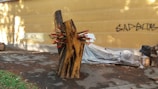 An abstract wooden sculpture consisting of two large wooden pieces with multiple red-tipped wooden spikes protruding from the center. The sculpture is placed on a paved area next to a yellow wall with graffiti. Various objects, possibly construction materials, are covered in plastic nearby.