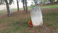 An old stone monument stands in a grassy area surrounded by trees. Inscriptions cover the face of the stone, and vibrant flowers are placed at its base. To the side, a historic stone wall stretches across the background.
