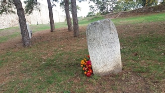 An old stone monument stands in a grassy area surrounded by trees. Inscriptions cover the face of the stone, and vibrant flowers are placed at its base. To the side, a historic stone wall stretches across the background.