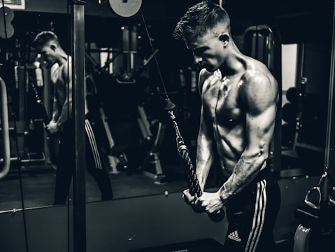 A muscular person is engaged in a workout at the gym, using a cable machine for arm exercises. The individual appears focused and determined, with a mirror reflecting their image in the background.