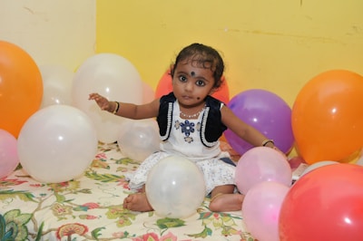 A young child sitting on a floral-patterned sheet surrounded by colorful balloons. The child is wearing a white dress with blue decorations and has dark hair with small accessories. Bright, mixed colored balloons in shades of orange, white, pink, and purple are positioned around the child.