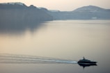 A serene seascape features a luxury yacht moving across calm waters with gentle ripples trailing behind. In the background, there are towering cliffs topped with small white structures, likely a coastal town, under a hazy sky.