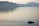 A serene seascape features a luxury yacht moving across calm waters with gentle ripples trailing behind. In the background, there are towering cliffs topped with small white structures, likely a coastal town, under a hazy sky.