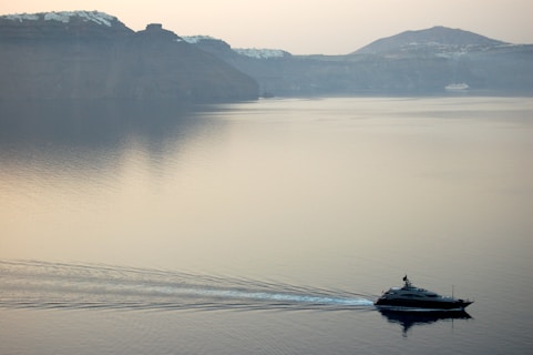 A serene seascape features a luxury yacht moving across calm waters with gentle ripples trailing behind. In the background, there are towering cliffs topped with small white structures, likely a coastal town, under a hazy sky.
