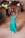 A young child in a vibrant green dress is joyfully dancing on a dirt floor. Her hands are raised, and her expression exudes happiness. In the background, a crowd of people are seated on wooden benches, watching the child's performance with interest.