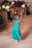 A young child in a vibrant green dress is joyfully dancing on a dirt floor. Her hands are raised, and her expression exudes happiness. In the background, a crowd of people are seated on wooden benches, watching the child's performance with interest.