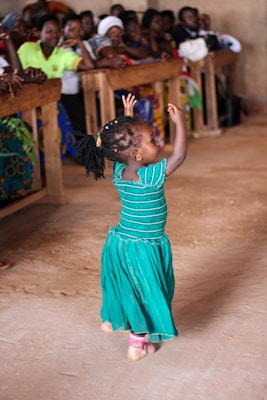 A young child in a vibrant green dress is joyfully dancing on a dirt floor. Her hands are raised, and her expression exudes happiness. In the background, a crowd of people are seated on wooden benches, watching the child's performance with interest.