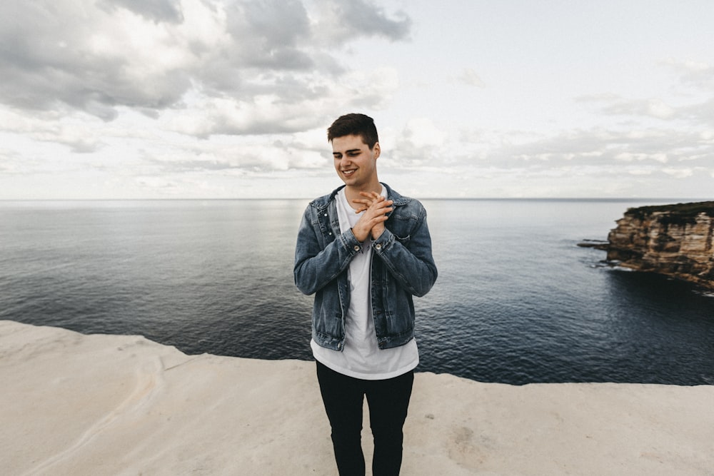 man in blue denim jacket standing on cliff near body of water