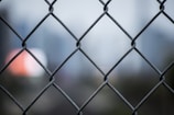 A chain-link fence in sharp focus with a blurred cityscape in the background. The foreground fence is metallic and forms a grid pattern, while the background shows out-of-focus elements, likely buildings and lights.