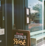A storefront with a large window displaying reflections and a sign advertising 'Dove's Summer of Rosé' outside. The window shows menus and interior elements of a diner environment, with reflections of nearby street scenes including a parked car and pedestrians.