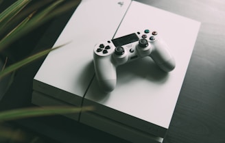 A white gaming console is placed on a dark surface, with a matching white controller resting on top. The console features smooth, clean lines and a sleek design. The surrounding area is slightly blurred, giving focus to the gaming equipment.
