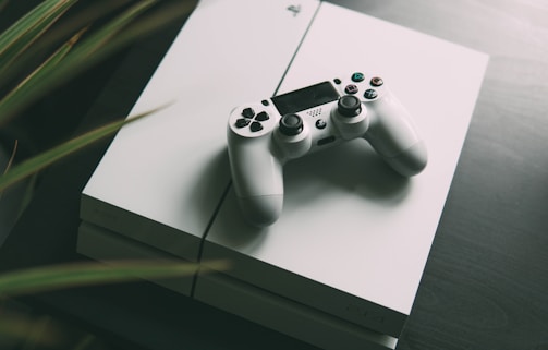 A white gaming console is placed on a dark surface, with a matching white controller resting on top. The console features smooth, clean lines and a sleek design. The surrounding area is slightly blurred, giving focus to the gaming equipment.