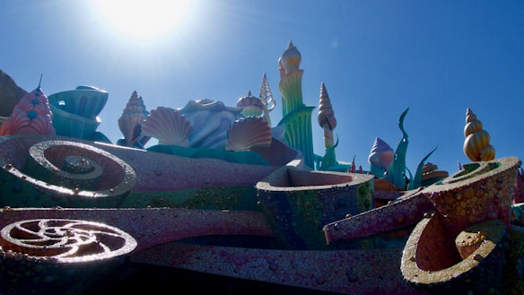 A whimsical, fantasy-themed structure adorned with oversized seashells and coral-like formations under a bright sky. The architecture resembles an underwater kingdom with vibrant, multicolored textures.