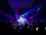 A vibrant concert stage illuminated with colorful lights, featuring a large display screen and numerous lighting rigs. There are silhouettes of people and equipment in the foreground, indicating a live performance setting. The interior architecture frames the scene, creating a sense of depth.