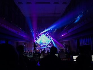 A vibrant concert stage illuminated with colorful lights, featuring a large display screen and numerous lighting rigs. There are silhouettes of people and equipment in the foreground, indicating a live performance setting. The interior architecture frames the scene, creating a sense of depth.