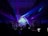 A vibrant concert stage illuminated with colorful lights, featuring a large display screen and numerous lighting rigs. There are silhouettes of people and equipment in the foreground, indicating a live performance setting. The interior architecture frames the scene, creating a sense of depth.