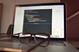 A laptop is open and displaying a programming interface with code written in a text editor. A pair of eyeglasses is placed on the keyboard in front of the laptop, creating a blur effect over the lower part of the screen.
