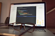 A laptop is open and displaying a programming interface with code written in a text editor. A pair of eyeglasses is placed on the keyboard in front of the laptop, creating a blur effect over the lower part of the screen.