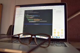 A laptop is open and displaying a programming interface with code written in a text editor. A pair of eyeglasses is placed on the keyboard in front of the laptop, creating a blur effect over the lower part of the screen.