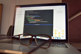 A laptop is open and displaying a programming interface with code written in a text editor. A pair of eyeglasses is placed on the keyboard in front of the laptop, creating a blur effect over the lower part of the screen.