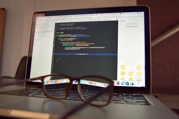 A laptop is open and displaying a programming interface with code written in a text editor. A pair of eyeglasses is placed on the keyboard in front of the laptop, creating a blur effect over the lower part of the screen.