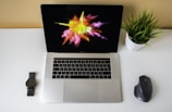 A modern workspace featuring an open laptop with a colorful abstract splash on the screen, a wristwatch on the left, a potted plant on the right, and a wireless mouse nearby, all placed on a white desk.