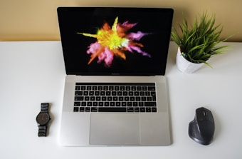 A modern workspace featuring an open laptop with a colorful abstract splash on the screen, a wristwatch on the left, a potted plant on the right, and a wireless mouse nearby, all placed on a white desk.