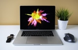 A laptop is placed on a white desk with a vibrant splash of yellow, pink, and orange colors displayed on its screen. To the left of the laptop is a black wristwatch and to the right is a black wireless mouse. A white pot containing a green plant with colored pencils is positioned beside the mouse. The background is a soft yellow wall.