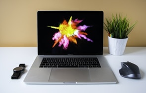 A laptop is placed on a white desk with a vibrant splash of yellow, pink, and orange colors displayed on its screen. To the left of the laptop is a black wristwatch and to the right is a black wireless mouse. A white pot containing a green plant with colored pencils is positioned beside the mouse. The background is a soft yellow wall.