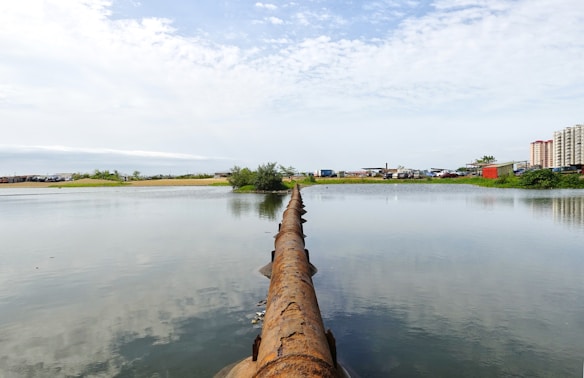 A large rusted pipeline extends across a calm body of water, dividing the scene with a clear reflection on the water's surface. In the background, there are green bushes and trees, with a mix of industrial structures and apartment buildings. The sky is mostly clear with scattered clouds.