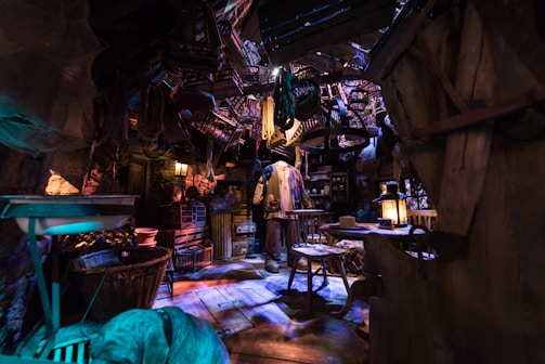 An intricately cluttered space resembling a workshop or storage room with dim lighting. Various objects hang from the ceiling, including tools and baskets. A figure stands in the center, surrounded by rustic wooden furniture and antique items like lanterns and crates, creating a mysterious, vintage atmosphere.