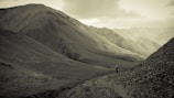 A person walks alone on a narrow path between towering mountains, surrounded by a vast and rugged landscape. The atmosphere is serene and the environment expansive.