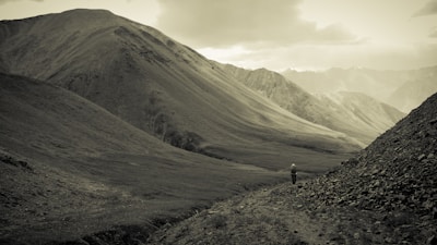 A person walks alone on a narrow path between towering mountains, surrounded by a vast and rugged landscape. The atmosphere is serene and the environment expansive.