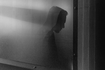 A shadowy silhouette of a person, likely a man, is visible through a misty glass, creating a mysterious and pensive atmosphere. The setting appears industrial or architectural with vertical lines on the right, possibly indicating a window or a partition.