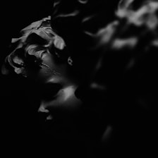 A highly stylized, monochrome image featuring a portion of a cherubic face. The play of light and shadow creates a dramatic and mysterious atmosphere. The texture and contours of the face are accentuated by the black and dark tones surrounding it.